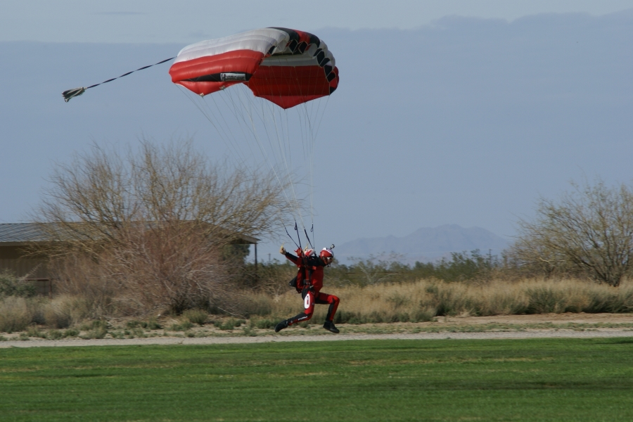 A parachutist with a red and white chute and matching jump suit starts a running motion just above the ground