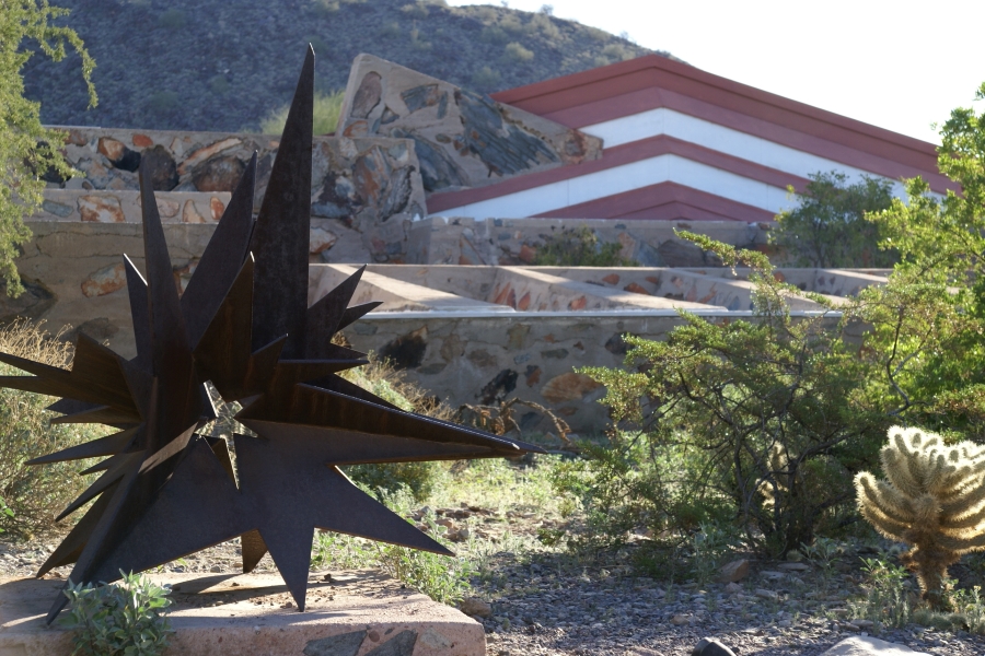 A spikey steel statue surrounded by scrub and cacti sits in front of low walls of large stones embedded in concrete
