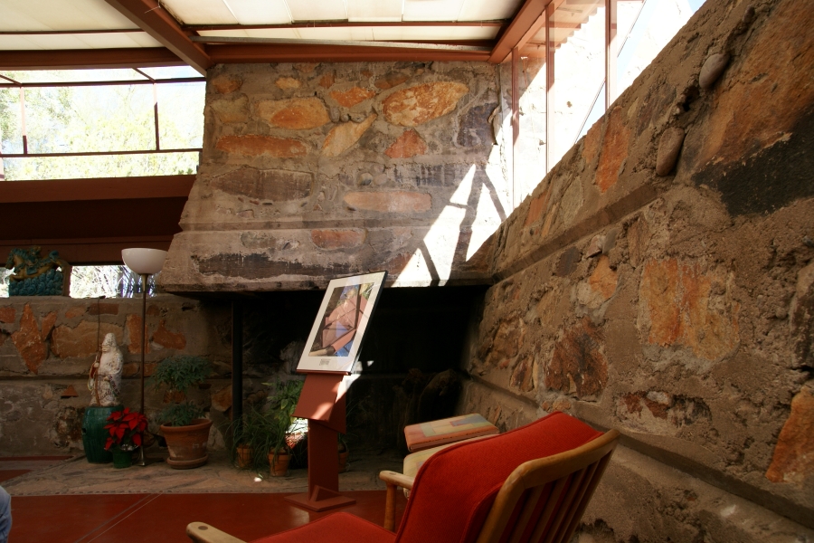 A large stone and concrete fireplace in the corner of a room with heavy walls with similar construction below strip windows letting in large amounts of natural light