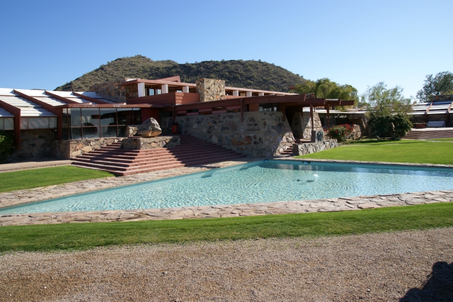 A shallow pool surrounded by manicured grass sits in front of a sprawling building of heavy stone and concrete walls with large horizontal beams making the building feel low to the ground