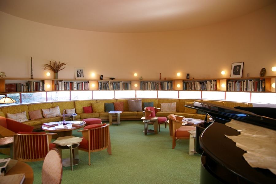 Inside a circular room a with a high ceiling and a continuous strip of windows between 4 foot and 5 foot 6 inches off the floor holds bench seating along the wall with bookshelves above the window and a piano along with several small table and chair seating groups