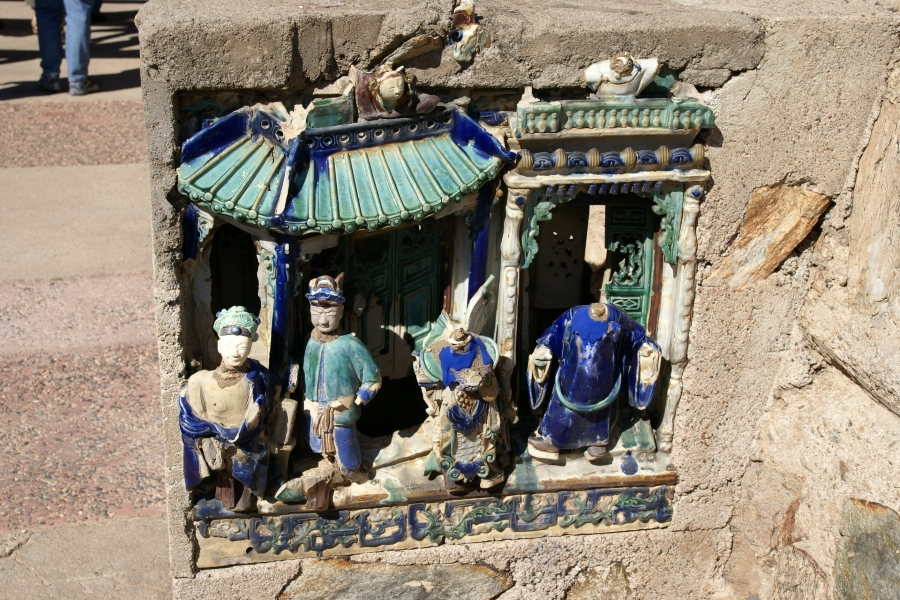 A faded blue and green porcelain diorama set in a concrete wall depicts Japanese figures in front of buildings