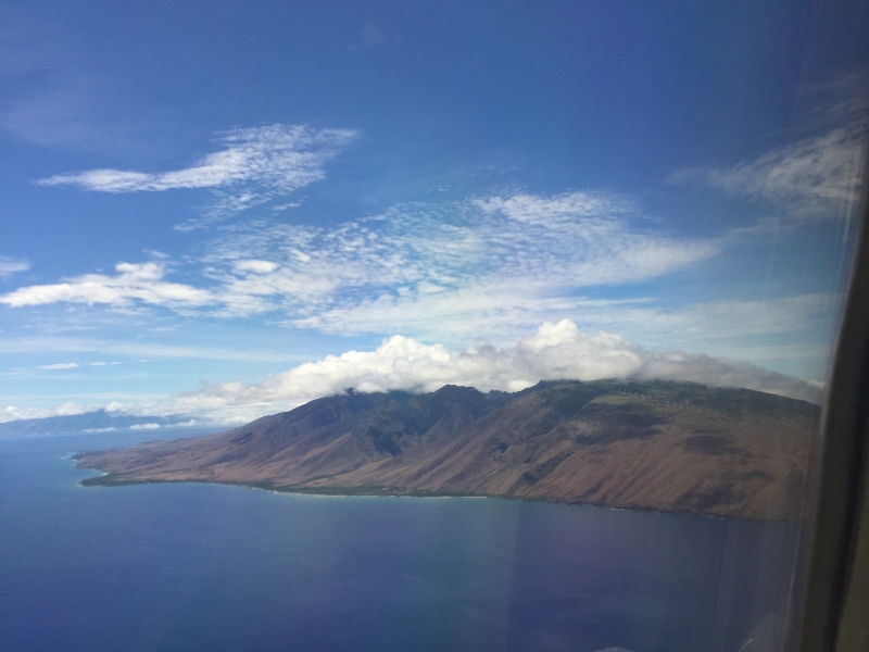The image shows a breathtaking aerial view of a mountainous island. The island's rugged terrain is sharply contrasted by the calm, blue ocean surrounding it. Wispy clouds drift across the sky, adding depth and dimension to the scene.