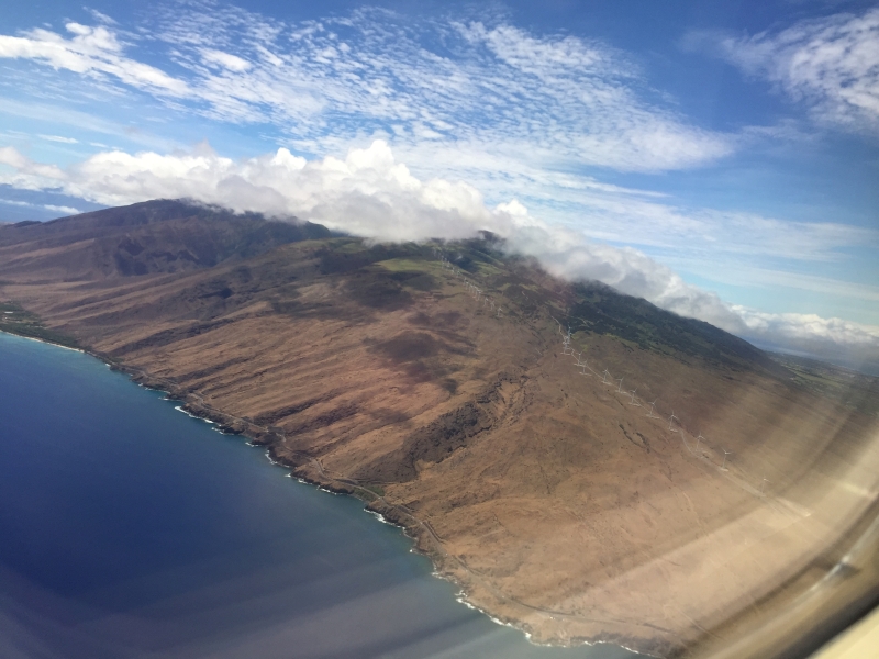 The image captures a bird's-eye view of a rugged island. A blue ocean abuts steep brown slopes, under a brilliant blue sky, dotted with fluffy white clouds.