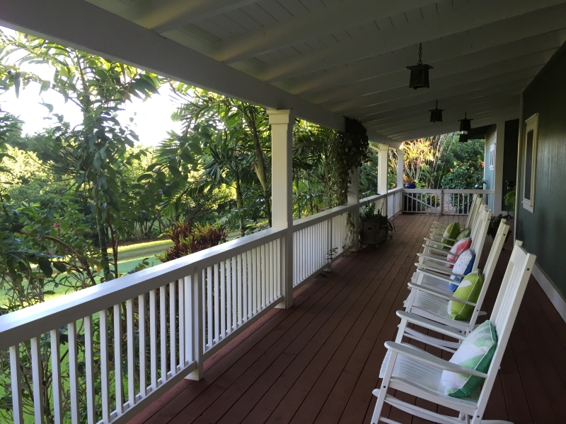 A spacious porch with a wooden floor and a white railing. Several rocking chairs with colorful cushions line the porch, inviting you to sit and relax. Beyond the railing, you can almost feel the cool breeze rustling through the lush green trees that stretch into the distance.