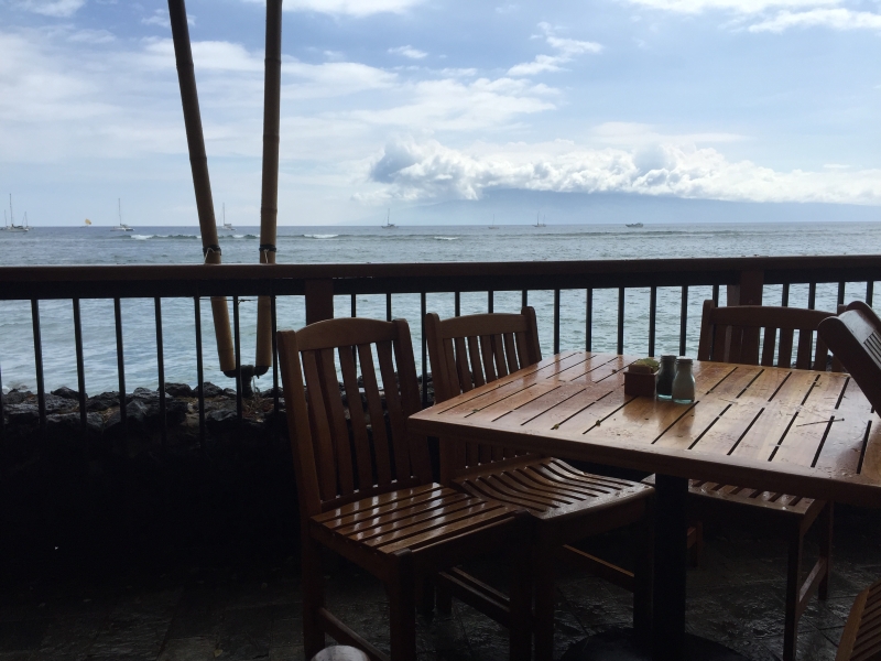 A wooden table and chairs set on a deck overlooking the ocean. Billiowing clouds in the distance, speak to cool ocean breeze.