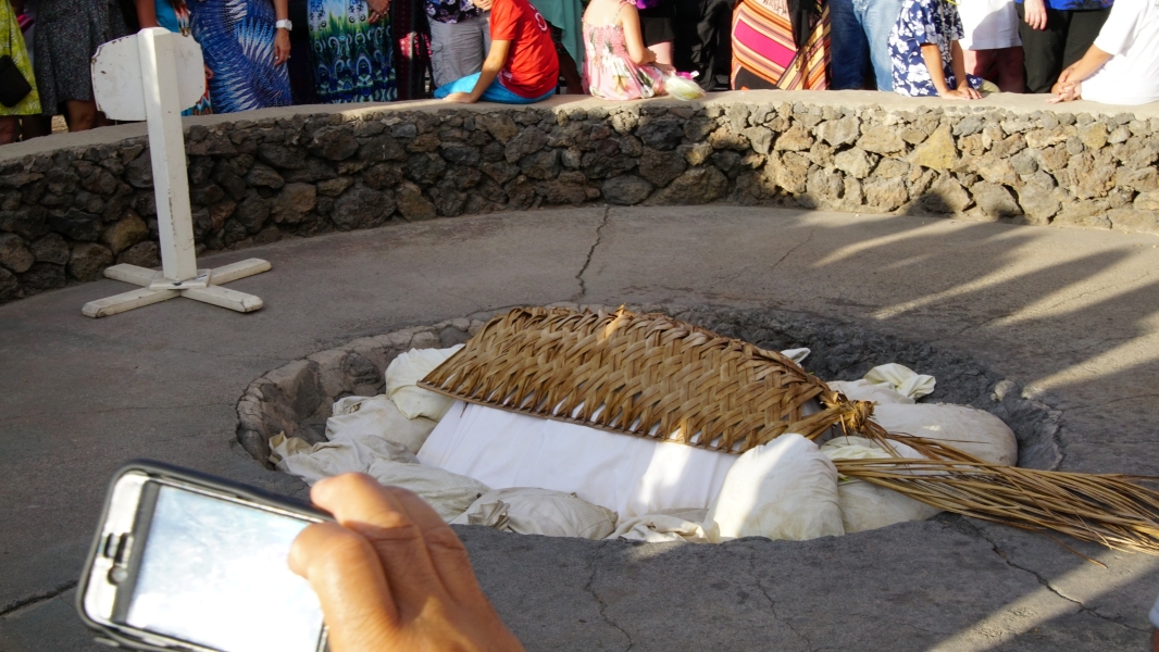 A circular pit, surrounded by a stone wall, holds a woven basket over a mound of white cloth. The cloth is surrounded by large white stones.