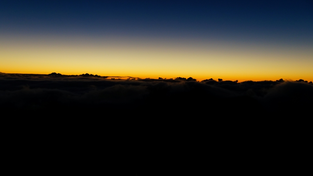 A breathtaking view of a sunrise from above the clouds. The sky is a canvas of vibrant colors, with deep blues blending into fiery oranges and yellows above a sea of fluffy clouds still shrouded in darkness.