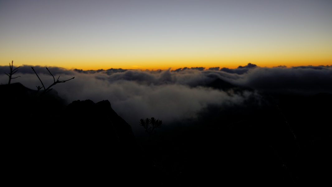 The sky is a canvas of vibrant colors, with pale blues blending into fiery oranges and yellows. Below, a sea of fluffy clouds appear as gray cushions on a dark landscape.