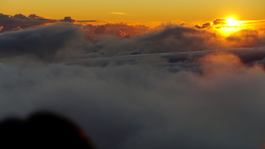The sun is a fiery ball of orange, casting a warm glow on the fluffy white clouds as it emerges over the horizon. The sky is a canvas of vibrant colors, with deep blues blending into fiery oranges and yellows. You can almost feel the warmth of the sun on your skin and the cool breeze on your face.