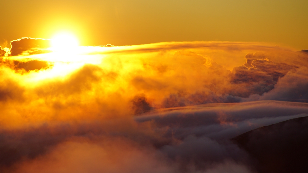 A breathtaking sunrise, with the sun casting a golden glow over a sea of clouds. The sky is ablaze with vibrant hues of orange, yellow, and pink, creating a mesmerizing spectacle. The clouds below look like fluffy cotton balls, their edges illuminated by the rising sun.