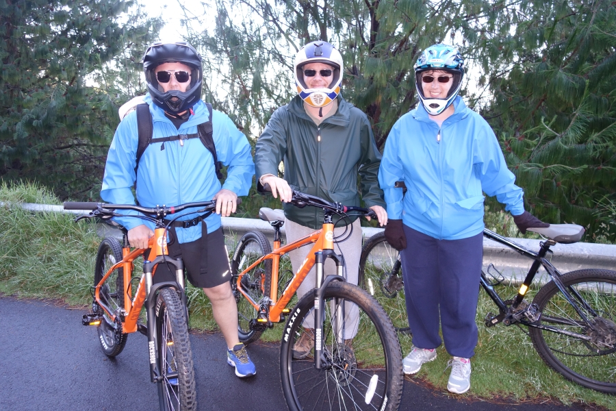 Three people stand next to their mountain bikes wearing blue windbreakers, sunglasses, and safety gear. The vibrant green background speaks to the tropical surroundings beyond the blacktop roadway.