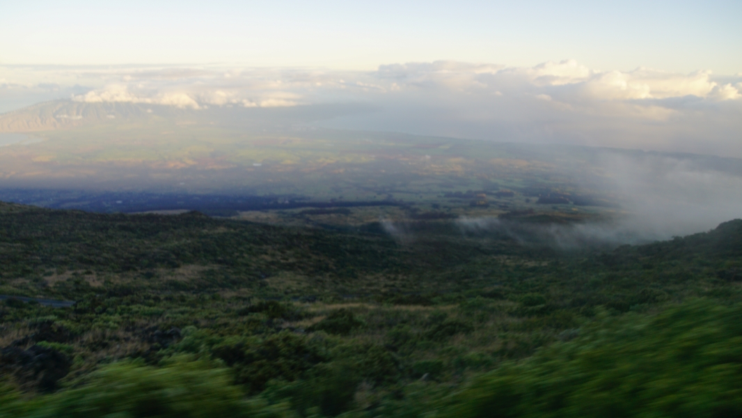 A misty morning view from a mountaintop. A blanket of fog softly covers the landscape below, obscuring the details of the rolling hills and distant coastline.
