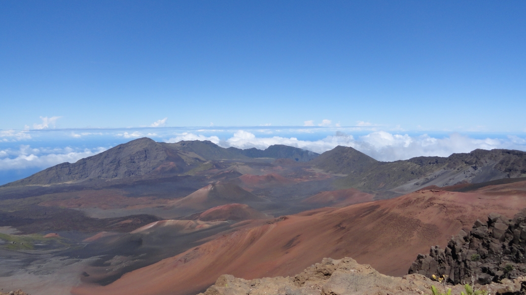 A vast, colorful landscape stretching out before you. A dormant volcano, its slopes a patchwork of red, brown, and black, dominates the scene. In the distance, the far side of the ancient caldera rises against a clear blue sky, punctuated by wisps of white clouds.