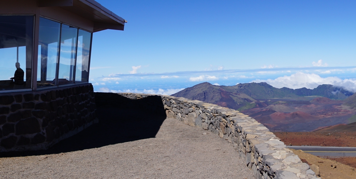 Imagine standing on a stone viewing platform, overlooking a breathtaking vista. A stone building with large windows frames the view, offering a glimpse of a distant mountain range shrouded in wispy clouds. Below, a winding trail snakes through a colorful landscape, hinting at the diverse terrain of this volcanic region.