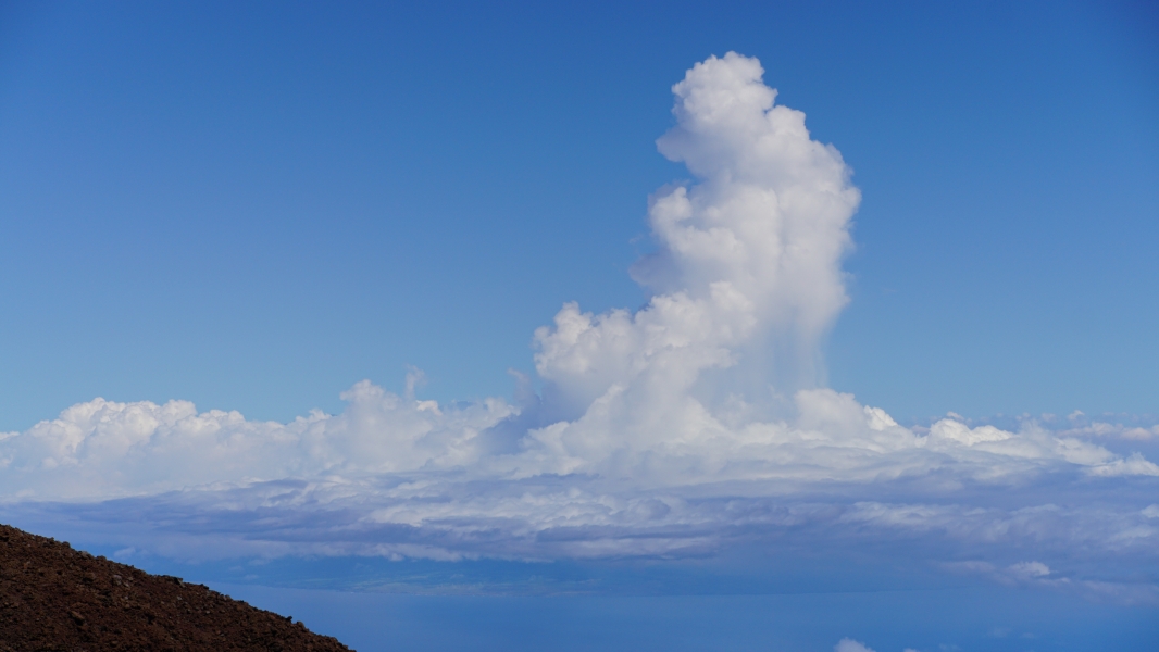 A towering cumulus cloud, shaped like a giant mushroom, rises against a bright blue sky. Below, a sea of fluffy white clouds stretches out towards the horizon above a mottled brown land mass, creating a breathtaking aerial view
