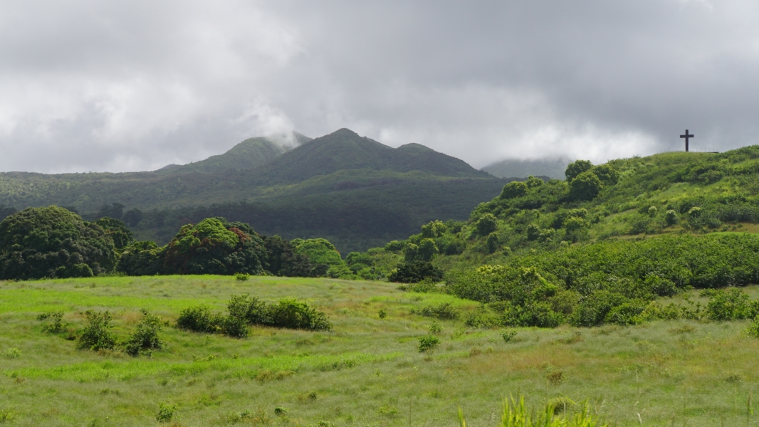 A serene landscape with rolling hills covered in lush green vegetation. In the distance, a mountain shrouded in mist rises majestically into the cloudy sky. Atop one of the hills, a simple wooden cross stands tall, adding a touch of spirituality to the scene.
