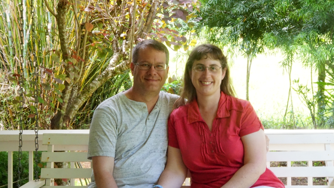 James and Julie sit side-by-side ona white porch swing with tropical flowering bushes in the background.