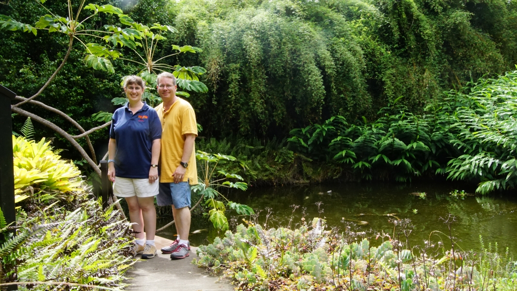 James and Julie stand on a narrow path with a small pond fed by a short waterfall surrounded by lush greenery.