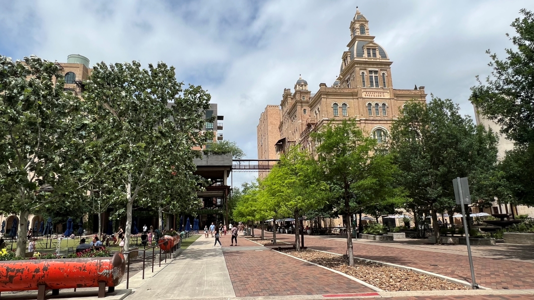 A rose-colored stone building rises above a park like area with plantings and trees around pedestrian a plaza