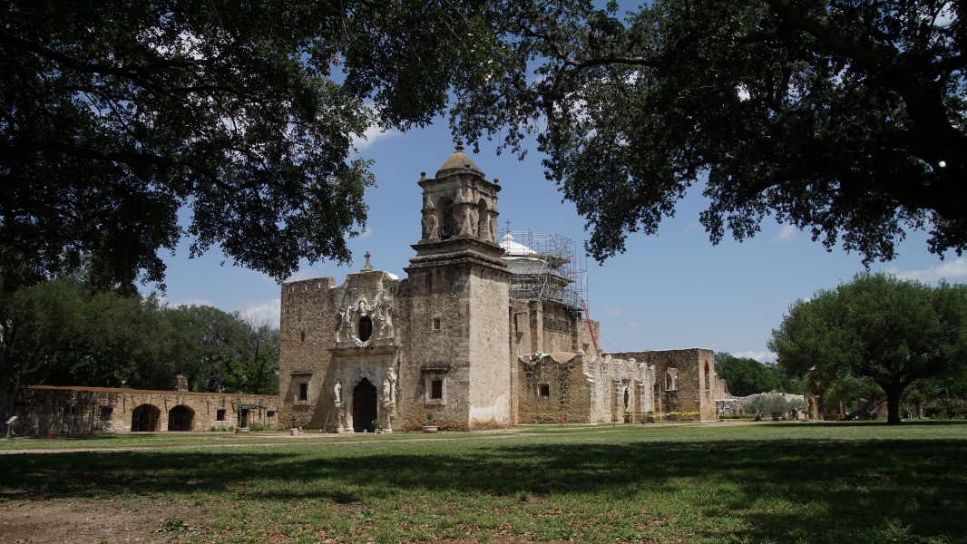 A white stone church with one tower with remnants of two-story stone buildings connected to the back stands beyond a green yard with trees spread around