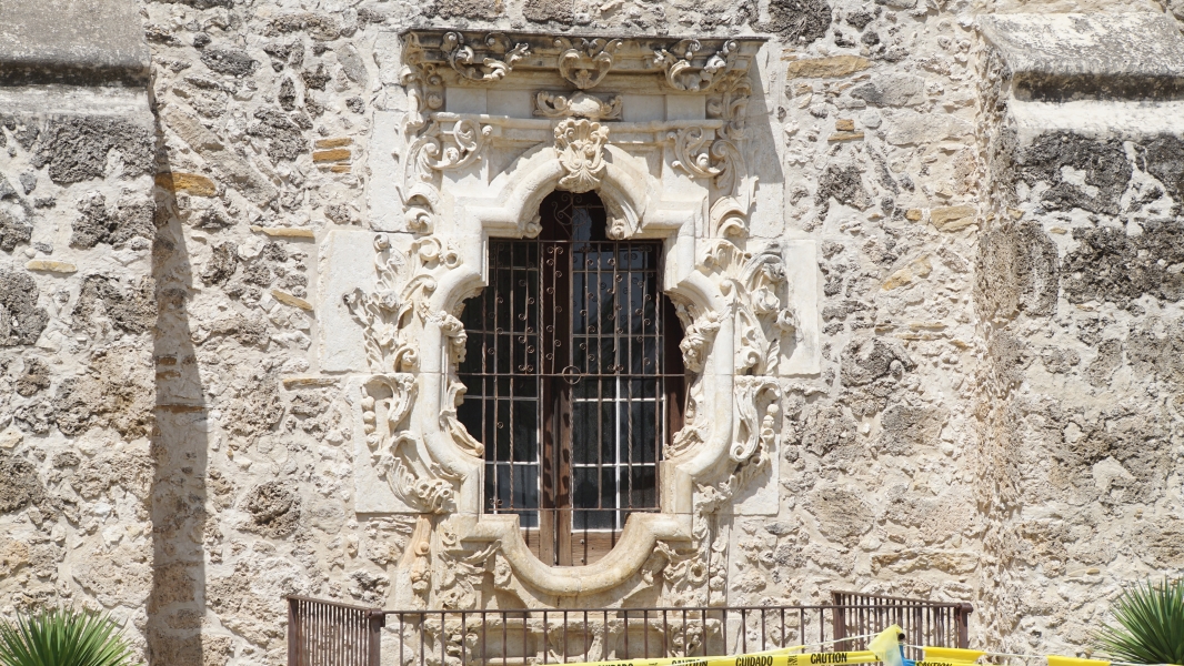 An ornately carved white stone window frame in a roughhewn stone wall surrounds an oval shaped window