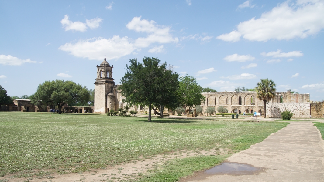 A path leads to the remains of a two-story structure attached to the back of a church passing an open field with sparsely spaced trees