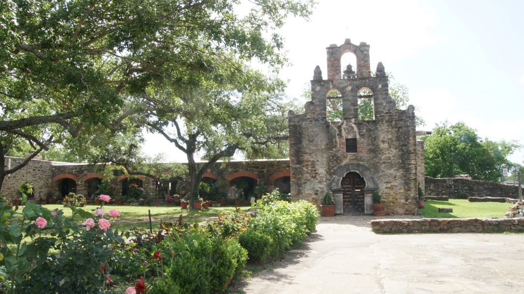 A stone church front with three bell opens in a flat wall with carefully maintained flowers along the path along the colonnade to church offices