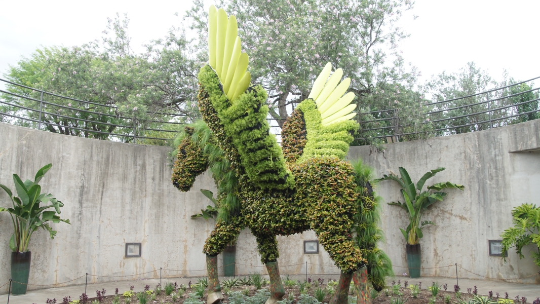 A large, winged horse made of various plants stands in a bed of flowers surrounded by a tall concrete wall