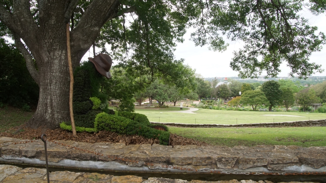 A large sleeping gardener made of various plants rests against a tree overlooking fields of grass