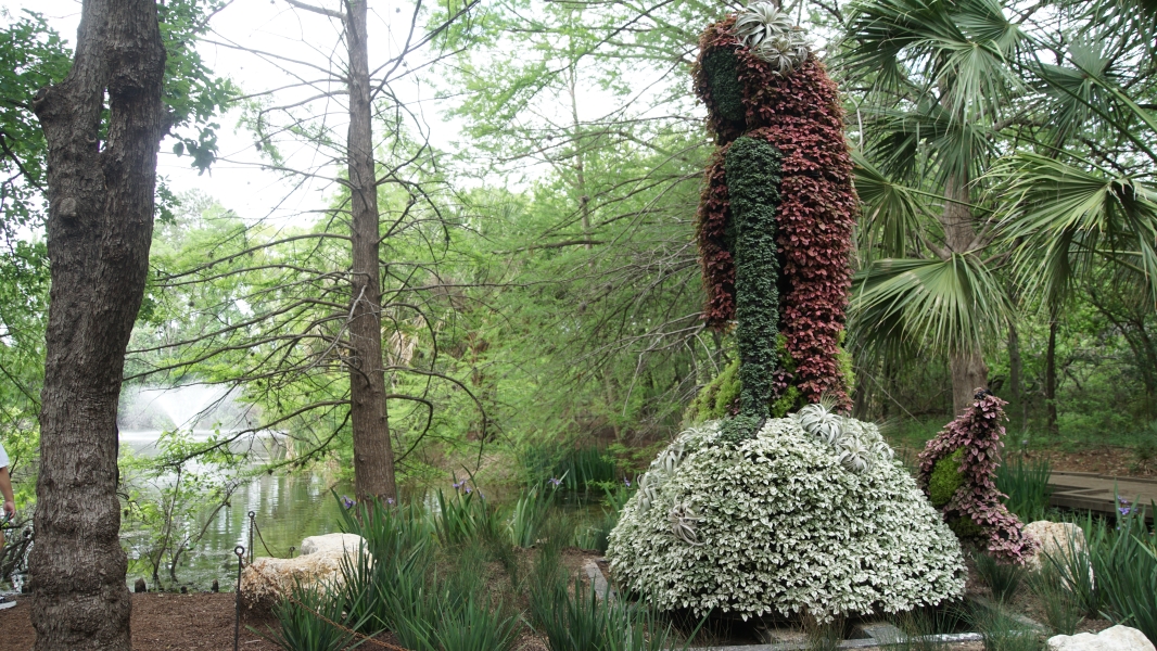 A nine-foot-tall mermaid made from white and pink flowers looks through the trees at a pond