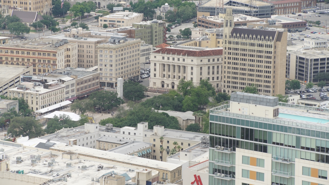 A white roofed cross shaped building is visible between the trees surrounded by taller buildings