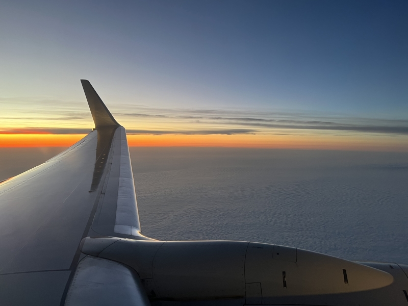 Bands of red and yellow fade into blues above a layer of white puffy clouds below an airplane wing