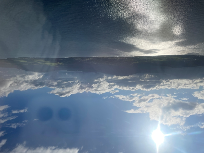 The sun reflects off the ocean in front of a green mountainous island with clouds around its peaks as seen outside an airplane window