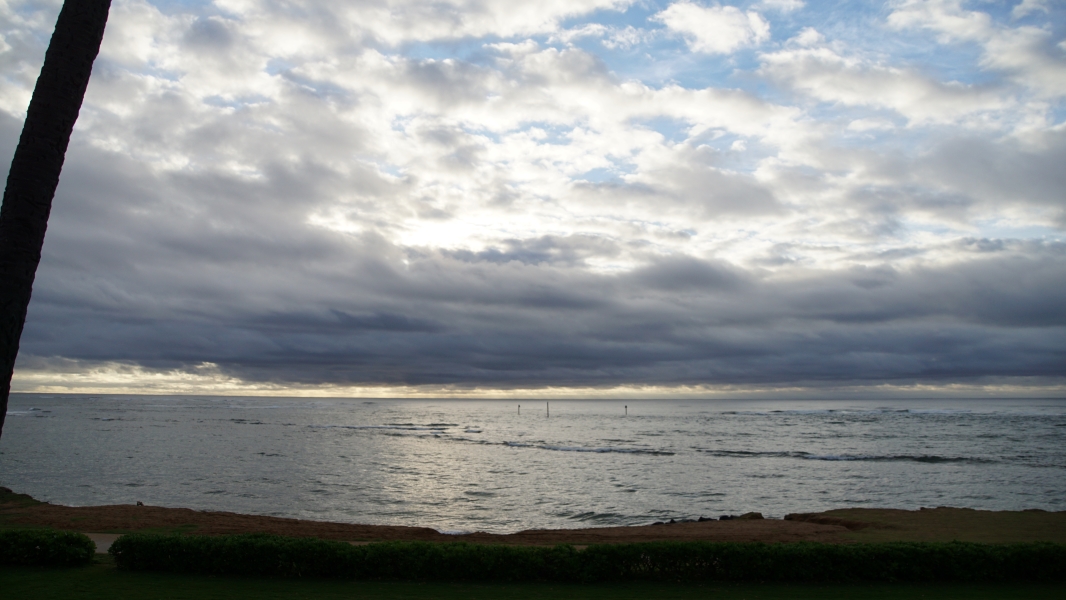 Broken clouds reveal patches of blue sky and yellow bands of sunshine above blue ocean waves and green grass