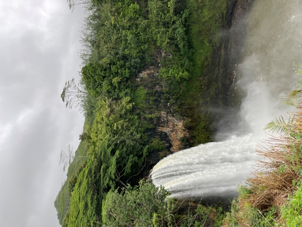 Mists rise from a lake surrounded by step stone cliffs covered with green plants with water waters of a wide waterfall cascade from a dark opening