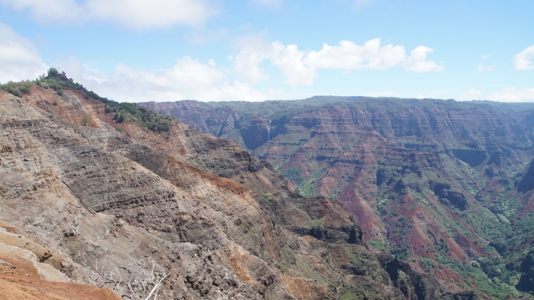 Blue skies with white puffy clouds over a deep canyon with lines of browns, grays, and green running horizontally across different crevices and rifts with a white water fall in the distance