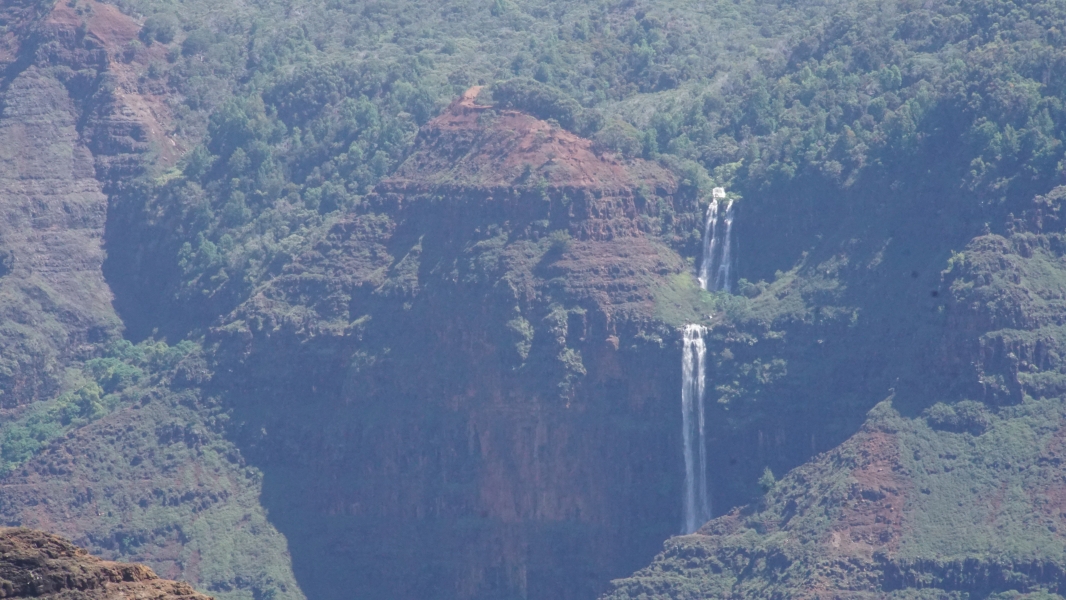 Green trees look like small shrubs compared to tall canyon walls near a two-level waterfall