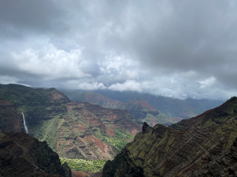 Dark clouds pass over allowing dappled sunlight to filter through onto a water fall and canyon walls of reds and browns with green scrub growing high and trees lower down