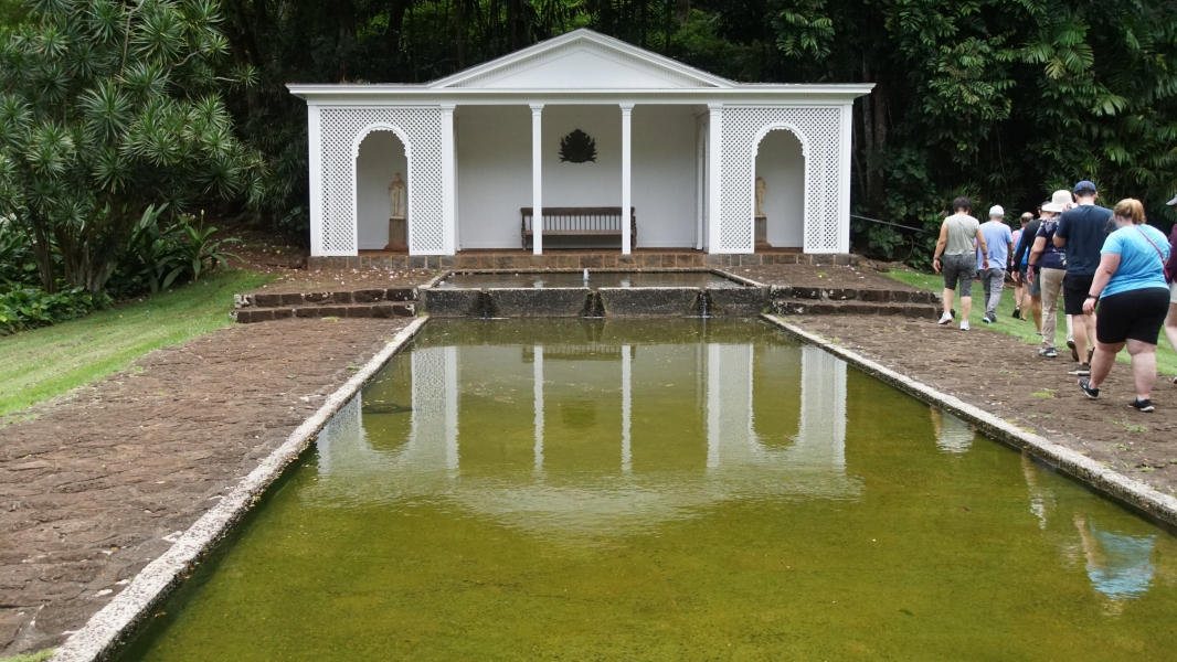 A white shade structure with Greek stylings stands at the end of a two-tier reflecting pool surrounded by a stone walkway, green lawn on a variety of tropical trees