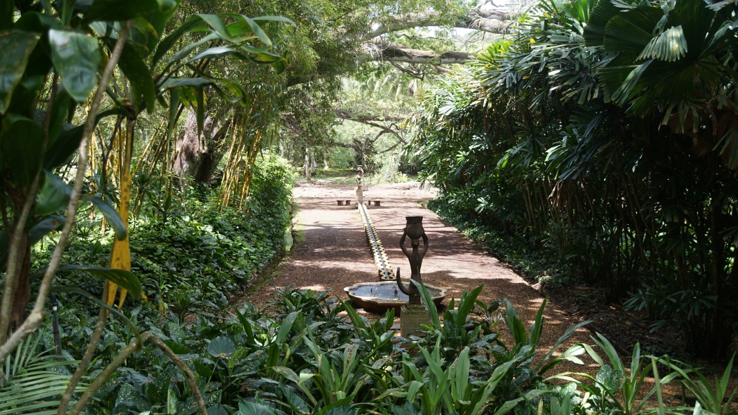Statues of mermaids stand at either end of a long stone water feature surrounded by leaver covered dirt with mid-height palms and bamboo running down each side