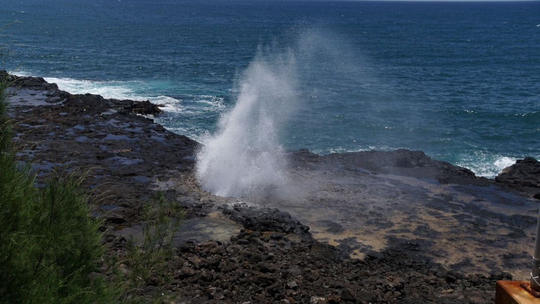 Water shoots vertically from a hole in black lava rock shelf above blue ocean waters with white foam breakers