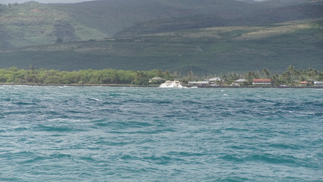 Choppy blue waters run to a black stone seawall where waves crash and spray extends as heigh as the eaves on a two-story building on the shore