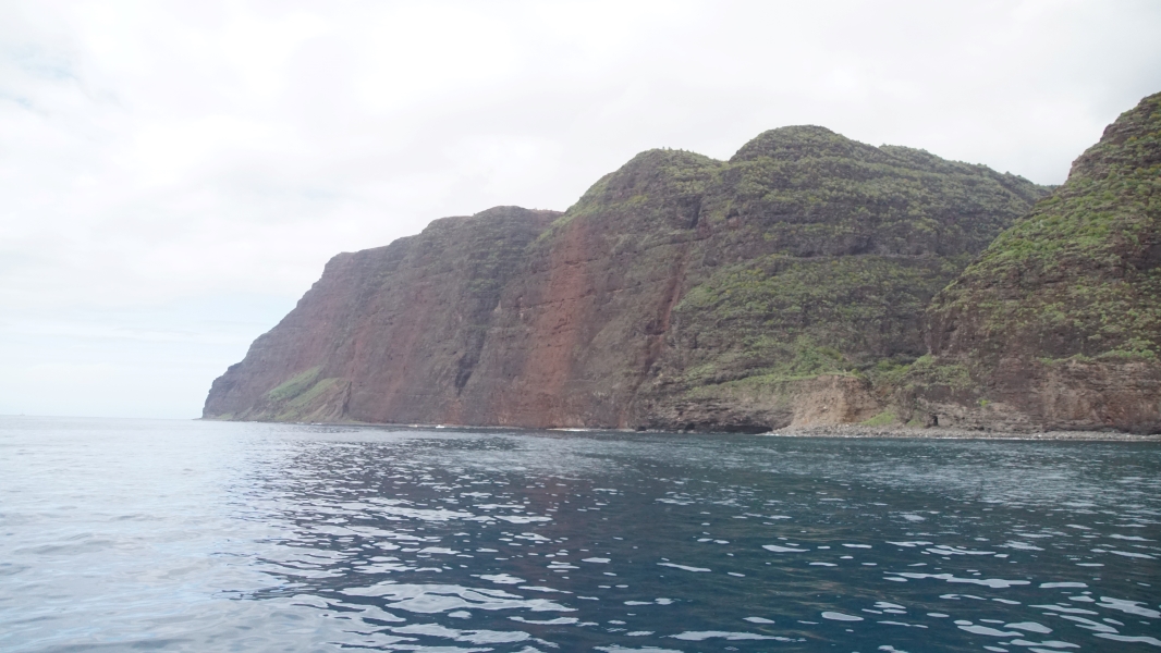 A line of shear red and black cliffs extends straight up over 1000 feet from blue ocean waters