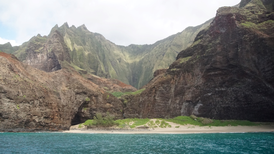 Shear walls several thousand feet tall covered in green scrub extend back behind a lower ridge above a sandy beach and blue water
