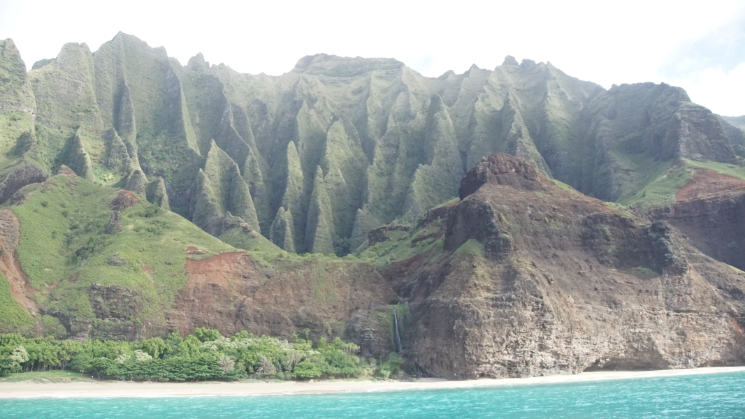 Green ridges twist and climb away from the water to a high crest reddish brown walls creating a ridge along a sandy beach