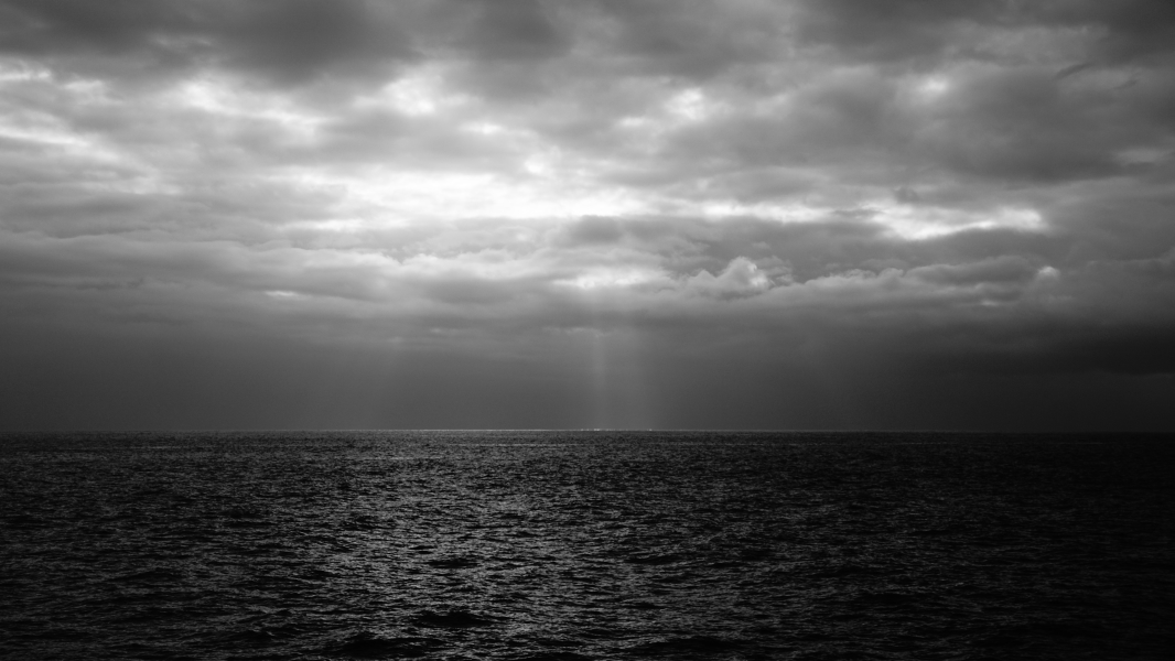 A black and white photo highlights the sun filtering through clouds and creating spotty rays dropping down to the water below