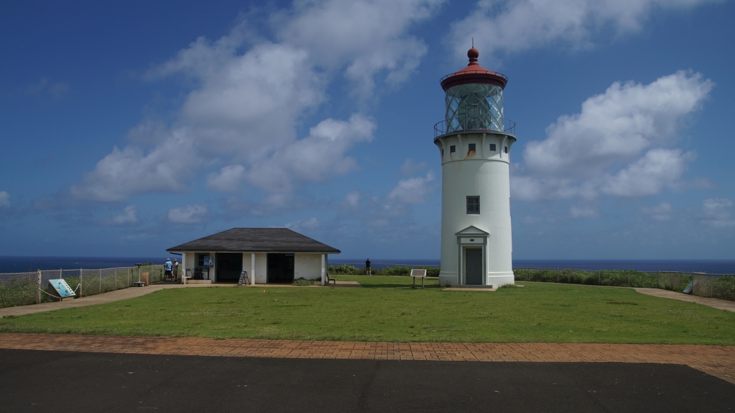 Brown pavers act as an edging between blacktop and a green grassy area with a small white building next to a three-story white lighthouse with a red roof on top below puffy clouds