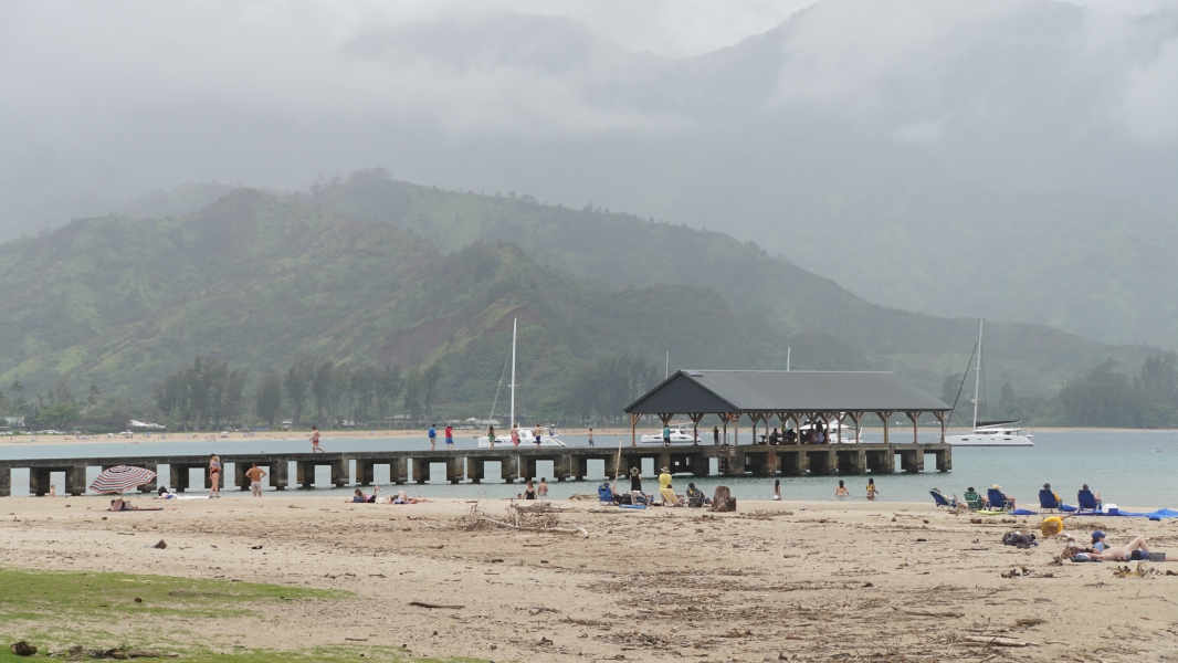 A low pier extends into the waters of a calm bay with beach goers in the foreground and cloud shrouded green hills in the distance