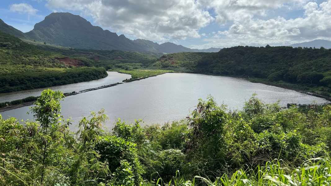 A river flowing around a bend towards a valley with steep walls on the left is partially separated from a large pond on the right by a low embankment
